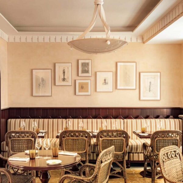 Restauracje otwarte w Niedzielę Wielkanocną 2025. Eleganckie wnętrze restauracji ukazuje dwa okrągłe stoliki z wiklinowymi krzesłami, jeden z resztkami posiłku i świecą. Wzdłuż ściany biegnie tapicerowana ławka z rzędem oprawionych grafik, a nad stolikami zwisa kremowy żyrandol. Całość tworzy przytulną i stylową atmosferę.