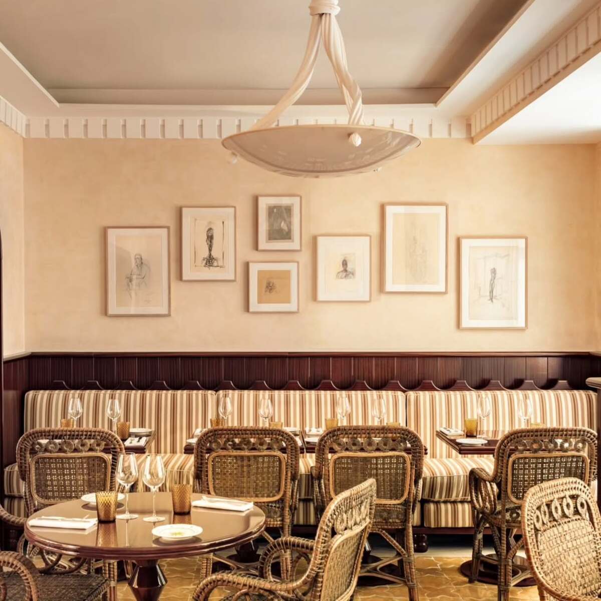 Restauracje otwarte w Niedzielę Wielkanocną 2025. Eleganckie wnętrze restauracji ukazuje dwa okrągłe stoliki z wiklinowymi krzesłami, jeden z resztkami posiłku i świecą. Wzdłuż ściany biegnie tapicerowana ławka z rzędem oprawionych grafik, a nad stolikami zwisa kremowy żyrandol. Całość tworzy przytulną i stylową atmosferę.