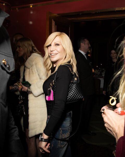 Donatella Versace kiedyś i dziś. Uśmiechnięta projektantka pozuje w czarnej bluzce na evencie. Na ramieniu ma zawieszoną czarną pikowaną torebkę.