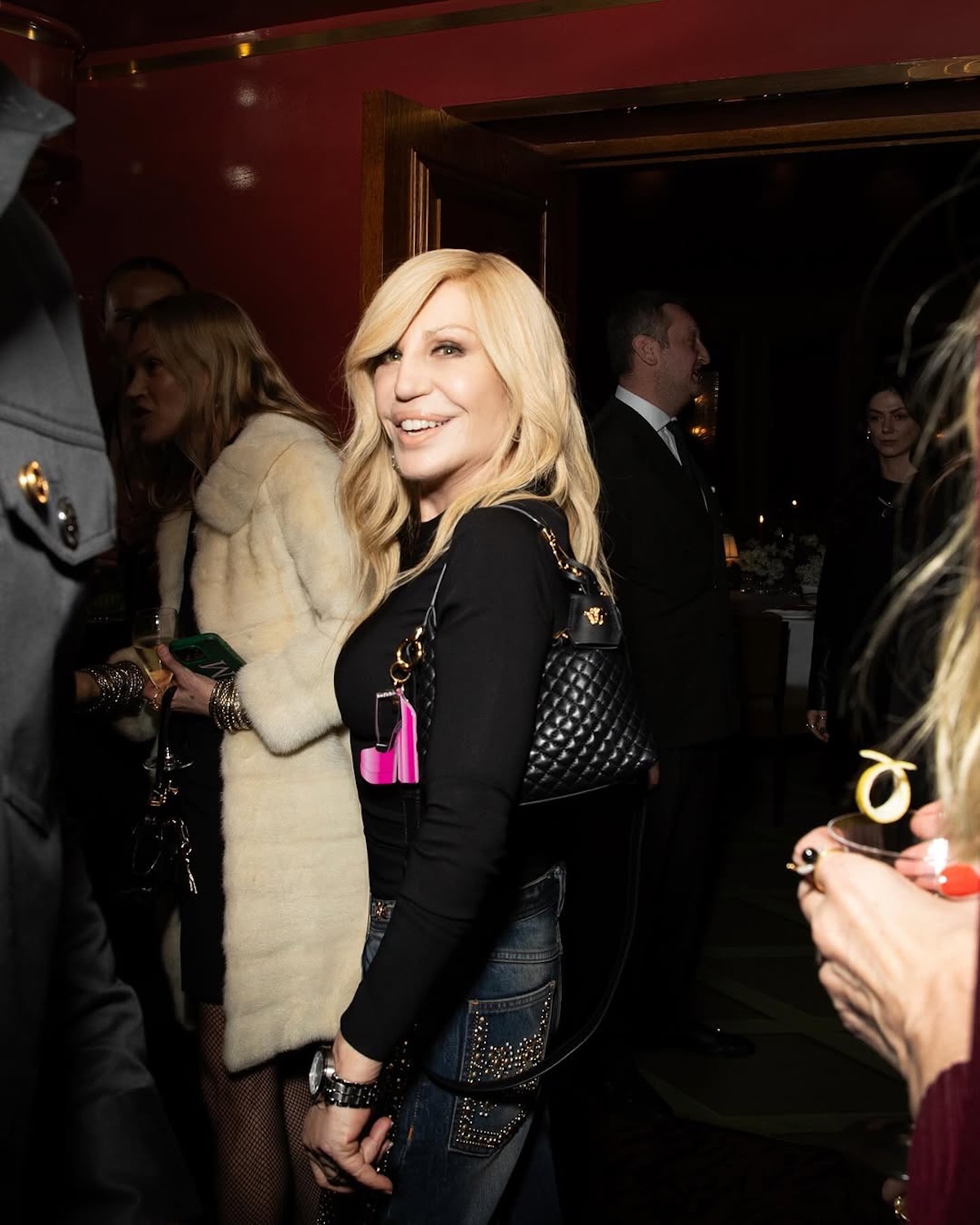 Donatella Versace kiedyś i dziś. Uśmiechnięta projektantka pozuje w czarnej bluzce na evencie. Na ramieniu ma zawieszoną czarną pikowaną torebkę.