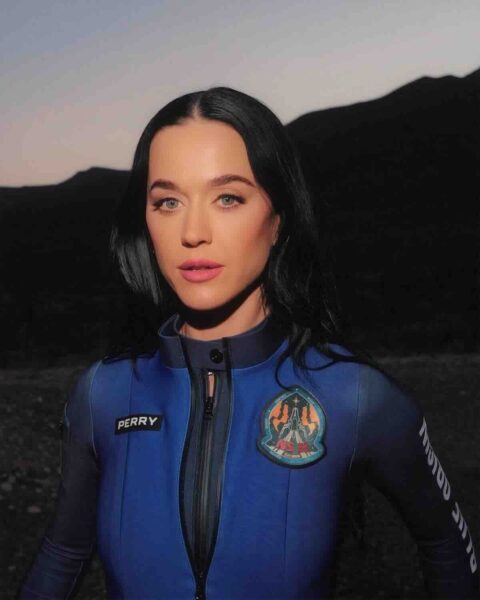 Katy Perry kosmos: Katy Perry stoi na tle zachodzącego słońca, opierając dłoń o twarz w zamyślonym geście. Ma na sobie kombinezon z logotypem Blue Origin, co symbolizuje jej udział w wyjątkowej kosmicznej misji złożonej wyłącznie z kobiet.