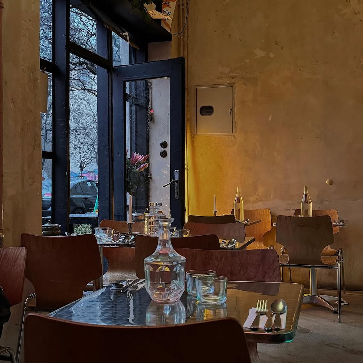 Wnętrze kameralnej restauracji z surowymi, nieotynkowanymi ścianami. Przy szklanych stołach ustawione są drewniane krzesła, na stołach stoją karafki z wodą, szklanki, sztućce oraz świece. Przez otwarte drzwi widać ulicę, samochody i drzewa bez liści w pochmurny dzień.