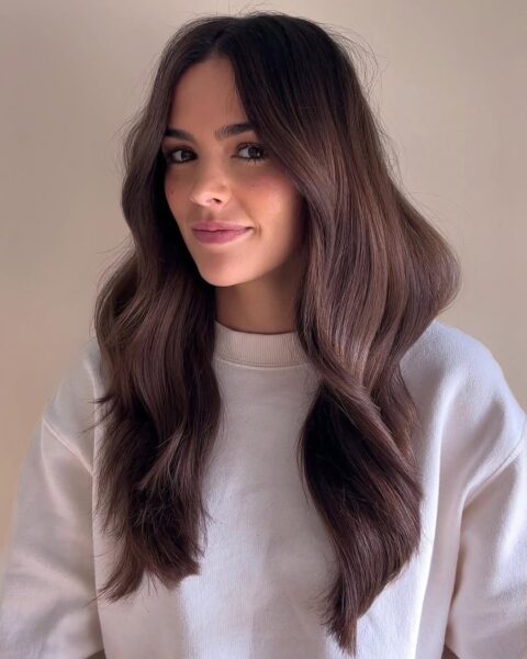 Młoda dziewczyna uśmiecha się delikatnie w szarej bluzie. Kobieta ma włosy w odcieniu Expensive Brunette – są rozpuszczone, błyszczące i lekko falowane.