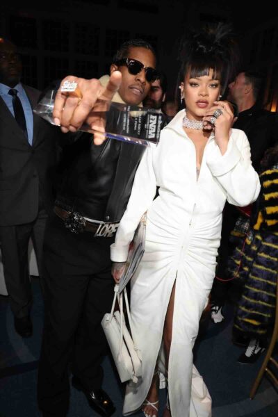 Najbardziej wpływowa para show biznesu: Rihanna w eleganckiej, białej sukni z marszczeniem i rozcięciem, obok A$AP Rocky’ego w czarnej skórzanej kurtce z napisem „LORD” i ciemnych okularach, trzymającego statuetkę FNA Footwear News Achievement Awards – para świętuje wspólny sukces.