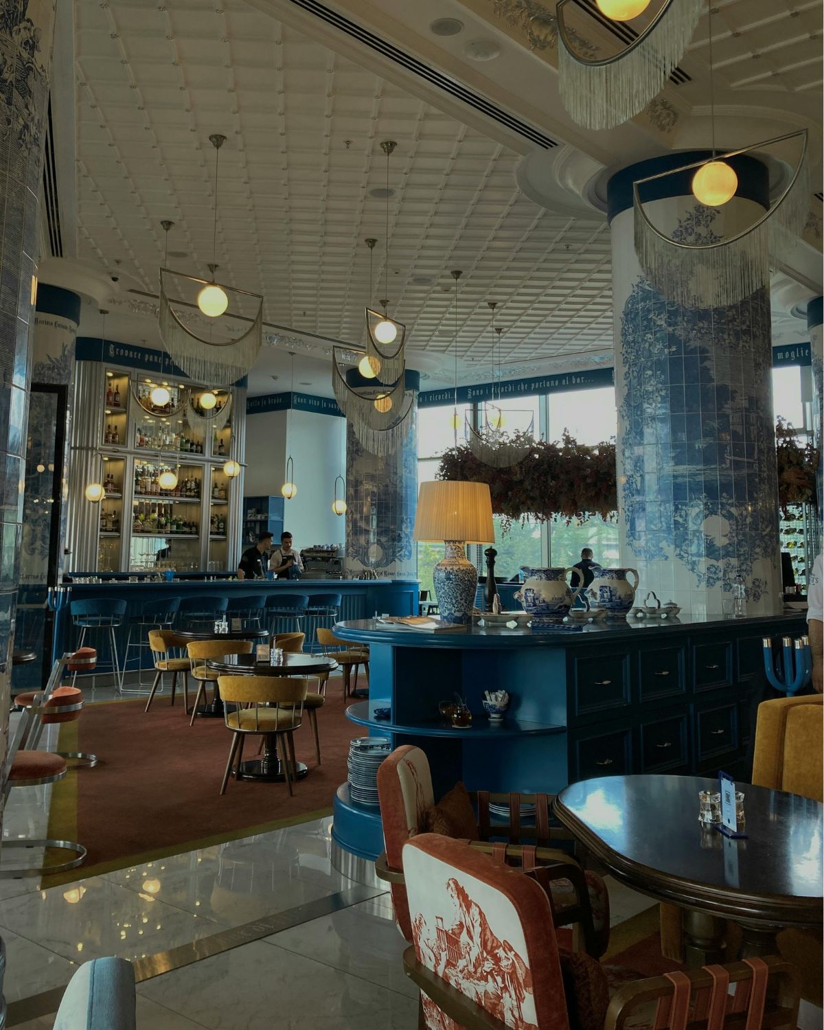 Zdjęcie, które przedstawia wnętrze restauracji, które wpisuje się w trend na restauracje z klimatem na majówkę w Warszawie: jest w kolorach niebieskim i bordo, z dekoracyjnymi kafelkami, kasetonami i sztukaterią oraz efektownym oświetleniem. Widać stoliki oraz bar restauracji.