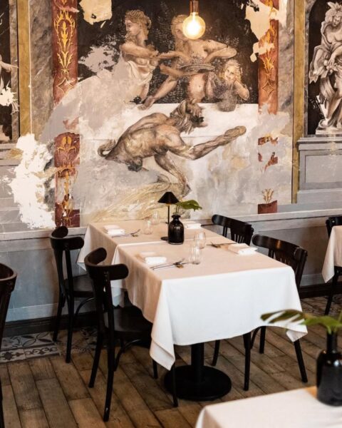 Zdjęcie, które przedstawia wnętrze restauracji La Luce. Widać stoliki nakryte białym obrusem i czarne krzesła ustawione pod ścianą z malowanymi freskami celowo postarzanymi, przypominającymi włoskie zabytki.