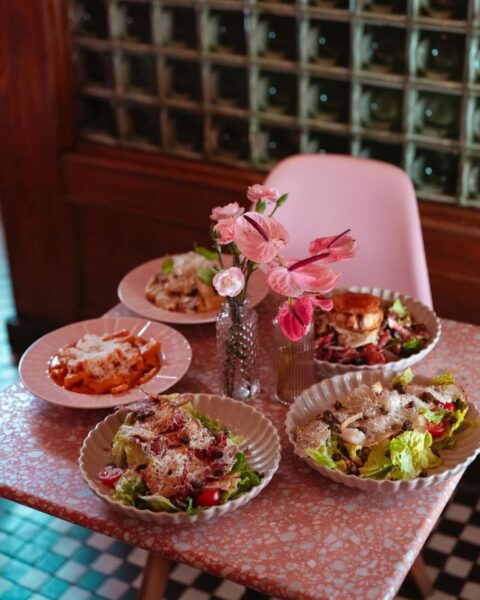 Zdjęcie, które przedstawia wnętrze restauracji Marcello. Widać różowy stolik z efektem lastryko, obok niego stoi różowe krzesło. W tle widać kafelkowaną w szachownicę podłogę, a także drewnianą ścianę z luksferami. Na stole stoją talerze z daniami i dwa małe wazony z kwiatami.