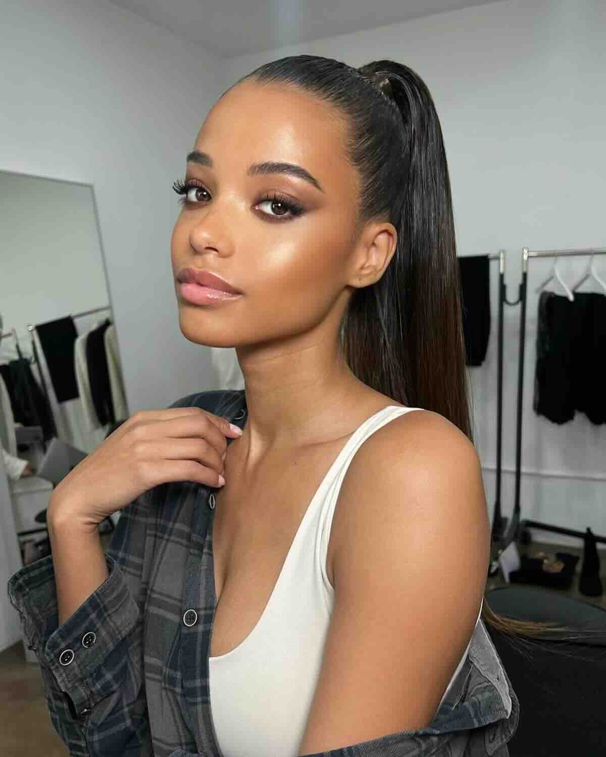 Ella Balinska kim jest?: Modelka w garderobie, z wysokim kucykiem i lśniącymi prostymi włosami, ubrana w biały top na ramiączkach i narzuconą luźno koszulę w kratę; jej makijaż podkreśla rozświetloną cerę i wyraziste oczy, a w tle widać wieszaki z ubraniami."