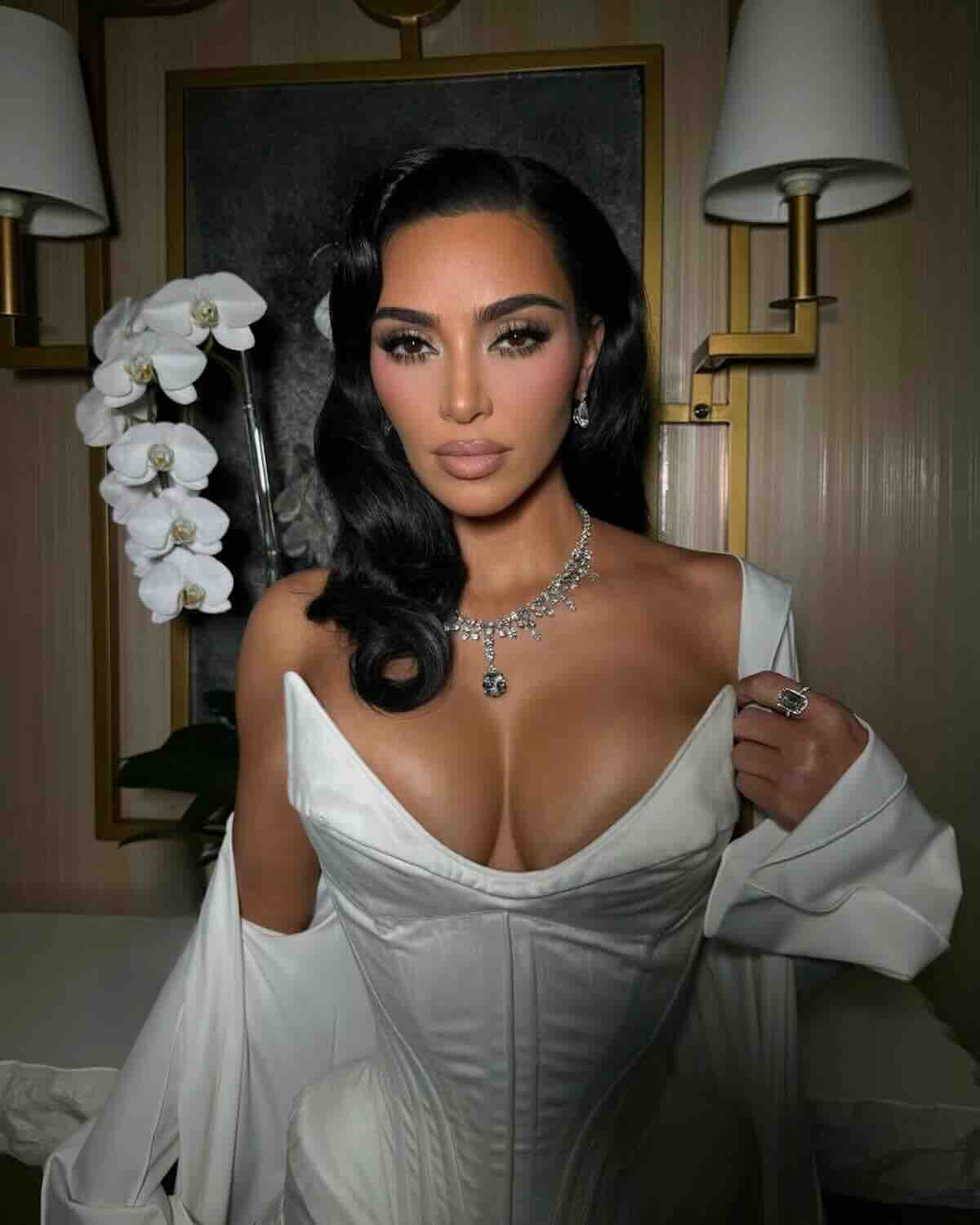 Kim Kardashian napad: Portret glamour kobiety w białym gorsecie i naramienniku, z diamentowym naszyjnikiem i makijażem w stylu "old Hollywood", na tle luksusowego wnętrza z orchideami.