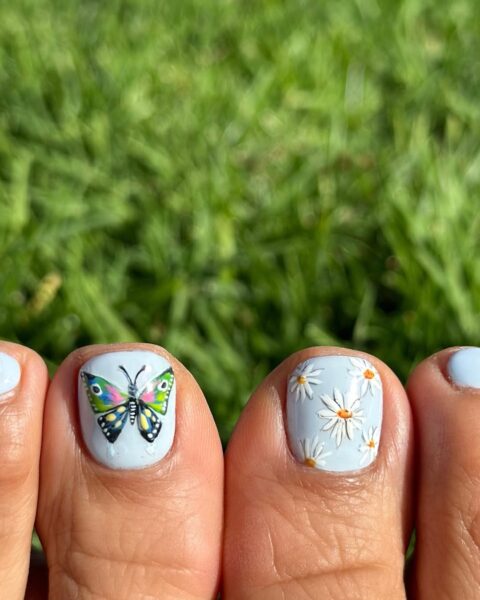 Uroczy, letni pedicure w pastelowych barwach – na błękitnym tle namalowany kolorowy motyl i stokrotki, wpisujące się w wakacyjne trendy na radosne, artystyczne zdobienia.