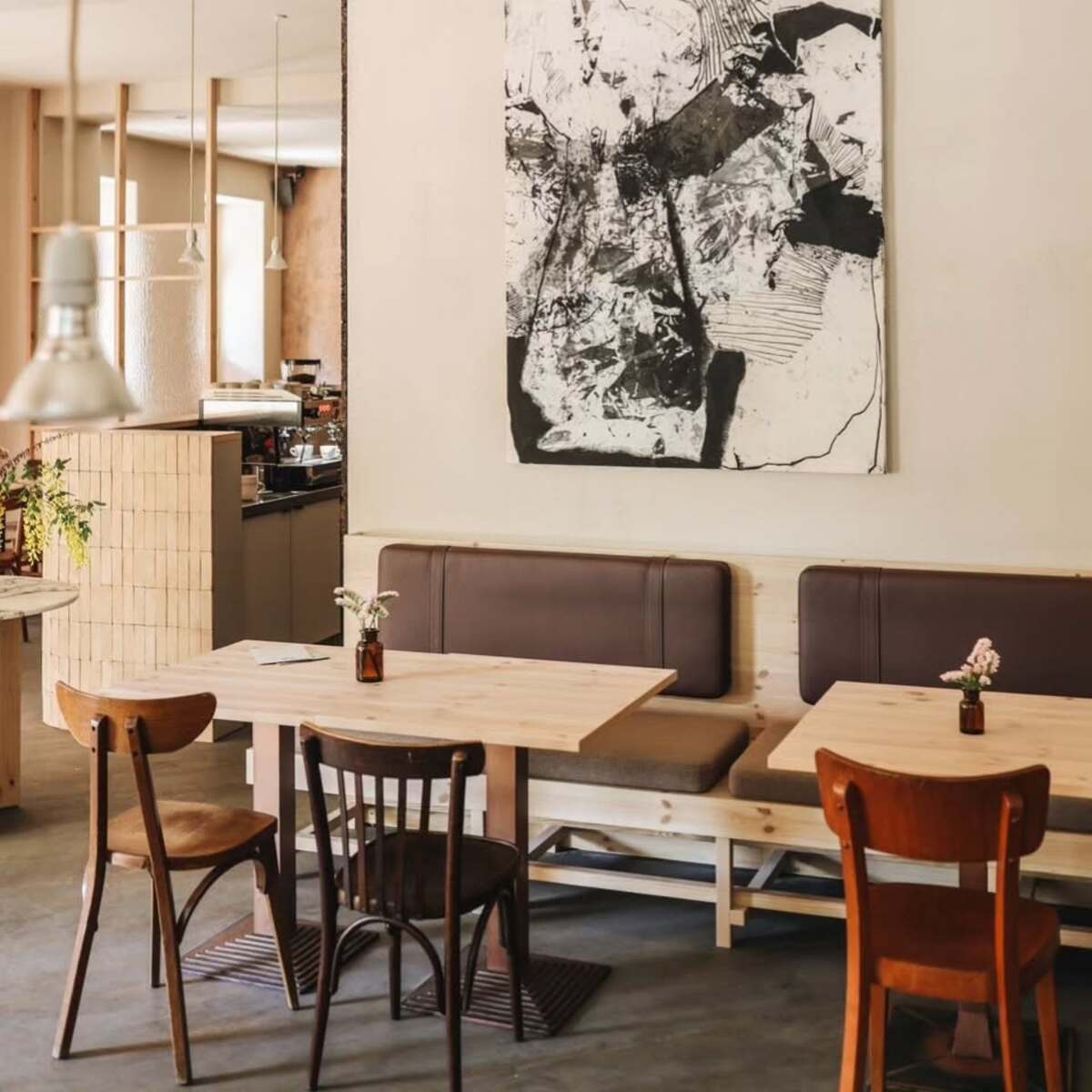 Restauracje otwarte w Poniedziałek Wielkanocny 2025 w Warszawie. Minimalistyczna restauracja z białymi ścianami, drewnianymi stołami i mieszanką różnych krzeseł. Dominuje czarno-biała abstrakcyjna grafika na ścianie.
