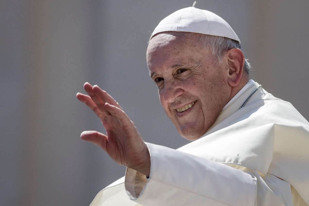 Na zdjęciu widać Papieża Franciszka, który macha do tłumu. Uśmiecha się szeroko, a jego postawa emanuje życzliwością i serdecznością. Jest ubrany w biały strój papieski, a na głowie ma tradycyjny papieski nakrycie głowy – zucchetto. W tle widoczny jest rozmyty, jasny krajobraz, który sugeruje, że zdjęcie zostało wykonane podczas jakiegoś publicznego wydarzenia.