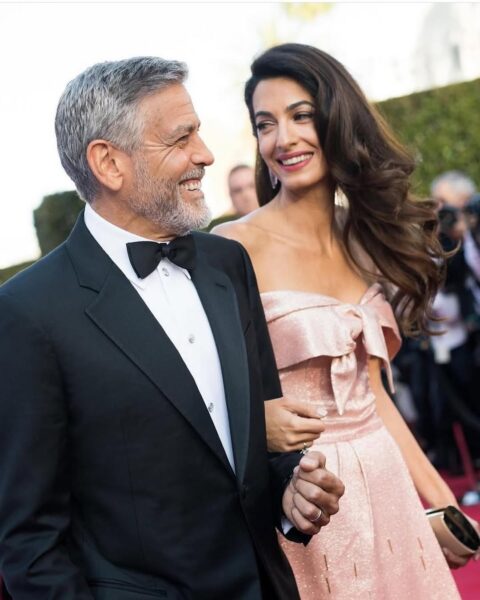Amal i George Clooney na czerwonym dywanie. George ma na sobie klasyczny czarny smoking z muszką, a Amal olśniewa w błyszczącej, różowej sukni z kokardą na biuście. Para uśmiecha się do siebie z czułością, idąc ramię w ramię.