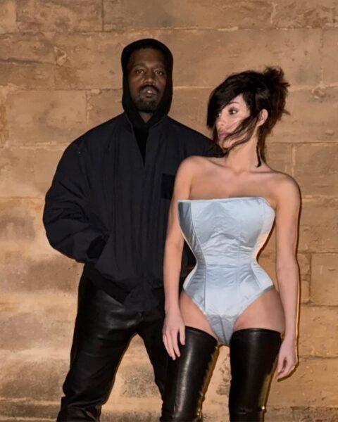 Zdjęcie Bianci Censori w srebrnym topie, wysokich, czarnych, lateksowych butach i kokiem. Za nią stoi Kanye West w kurtce czarnej bomberce, czarnych skórzanych spodniach i kapturem na głowie.