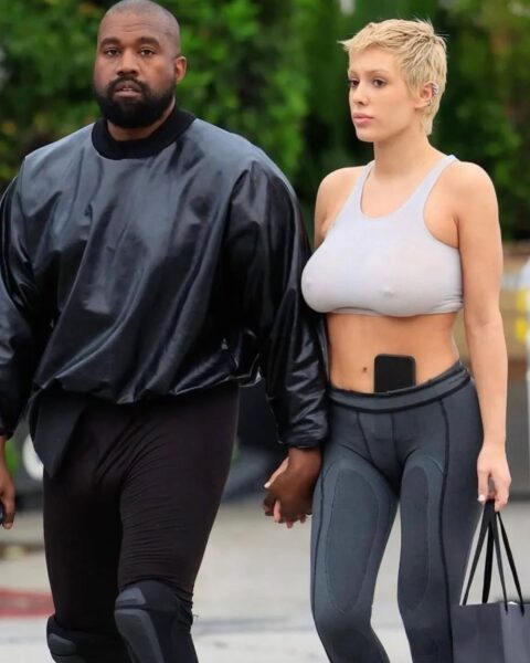 Zdjęcie Bianci Censori w kremowym topie i szarych legginsach. W pasie ma komórkę, a w ręce czarną torebkę. Ma krótkie, blond włosy. Idzie za rękę z Kanye Westem ubranym w czarne legginsy i czarną, skórzaną bluzę.
