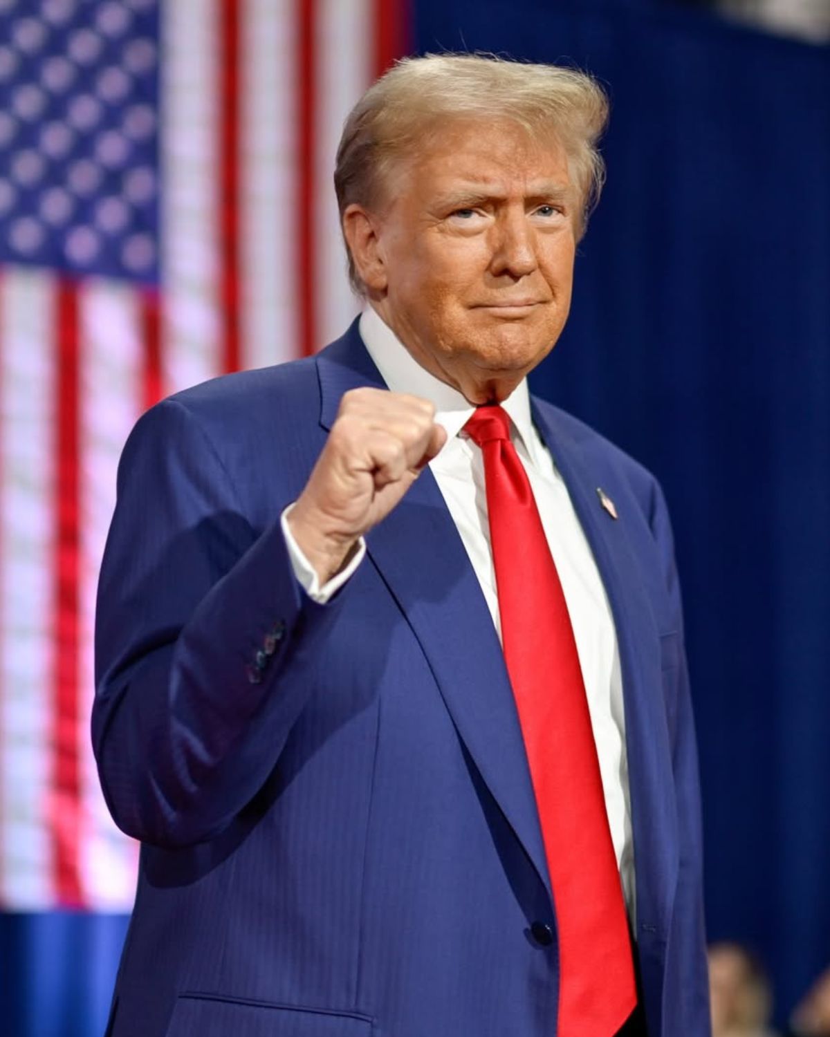 Donald Trump śledztwo 2025: zdjęcie prezydenta podczas wiecu wyborczego. Ubrany jest w granatowy garnitur, biała koszulę i czerwony krawat. Pozuje na tle flagi USA. Unosi pięść.