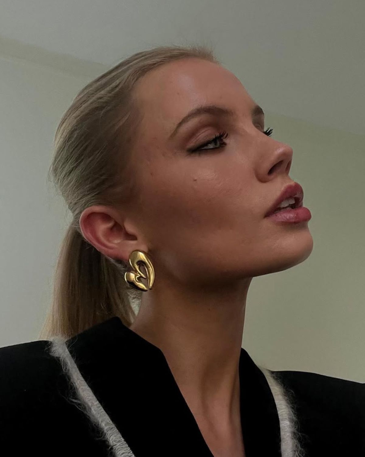 Zdjęcie kobiety, blondynki z kucykiem. Ma pomalowane oczy, ale z rzęsami ghost lashes, czyli prawie niewidocznymi. W uszach ma złote kolczyki w kształcie serc. Ubrana jest w czarną marynarkę.
