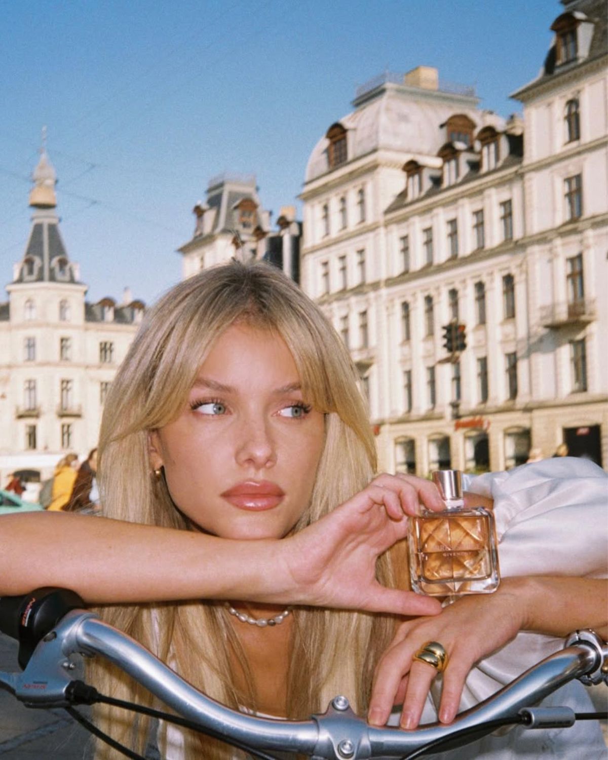 Zdjęcie kobiety, blondynki w rozpuszczonych włosach. Opiera się o kierownicę roweru i trzyma w ręce perfumy z nutą konwalii. Patrzy w bok. Widać naszyjnik z pereł na szyi oraz masywny, złoty pierścionek na środkowym palcu lewej ręki. W tle widać ulice Paryża.