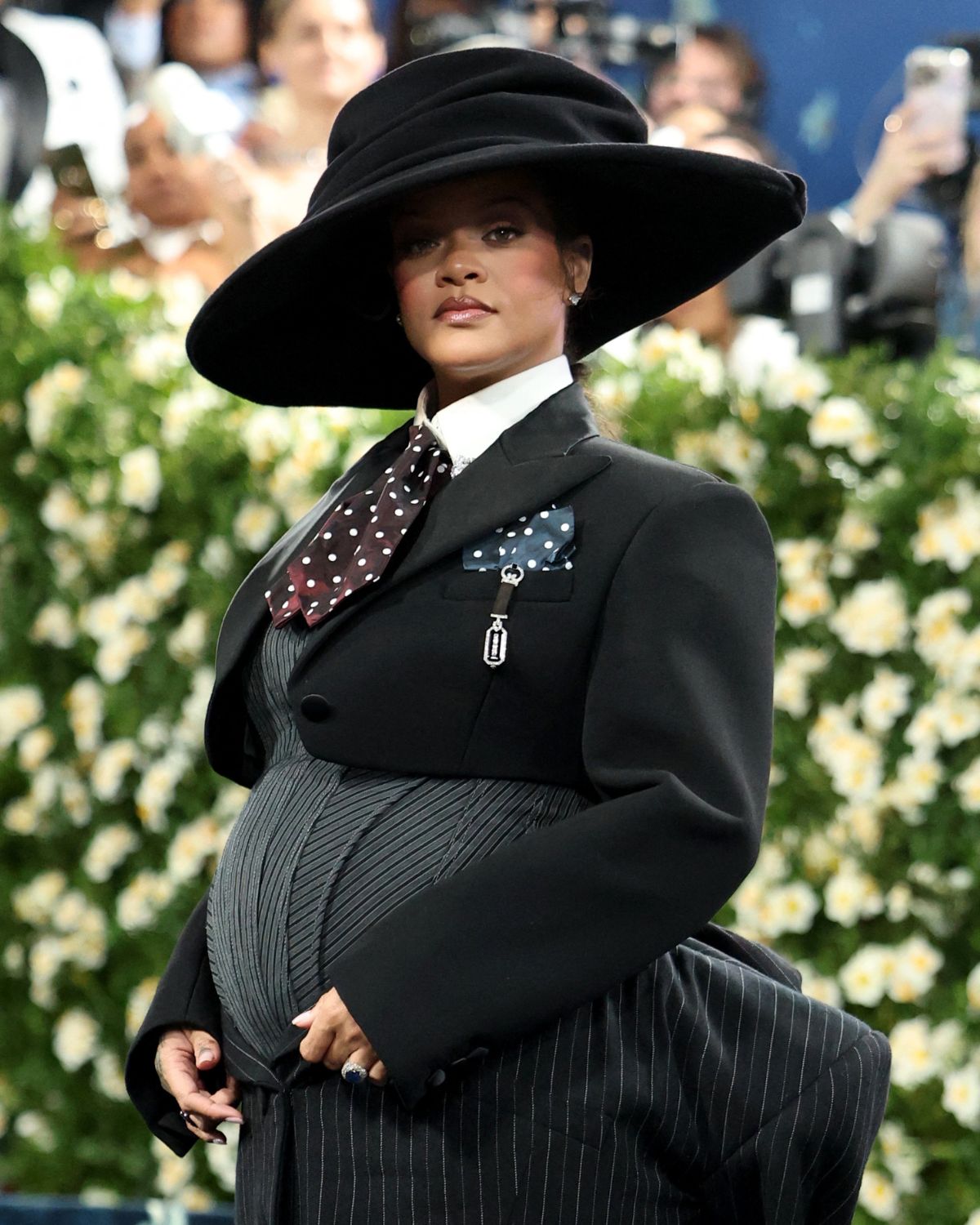 Rihanna i dzieci: ile gwiazda ich ma? Na zdjęciu widać Rihannę na Met Gala 2025. Ubrana jest czarną sukienkę z gorsetem, marynarką, koszulą oraz krawatem i kapeluszem na głowie. Za nią widać ogrodzenie z kwiatów i fotoreporterów.