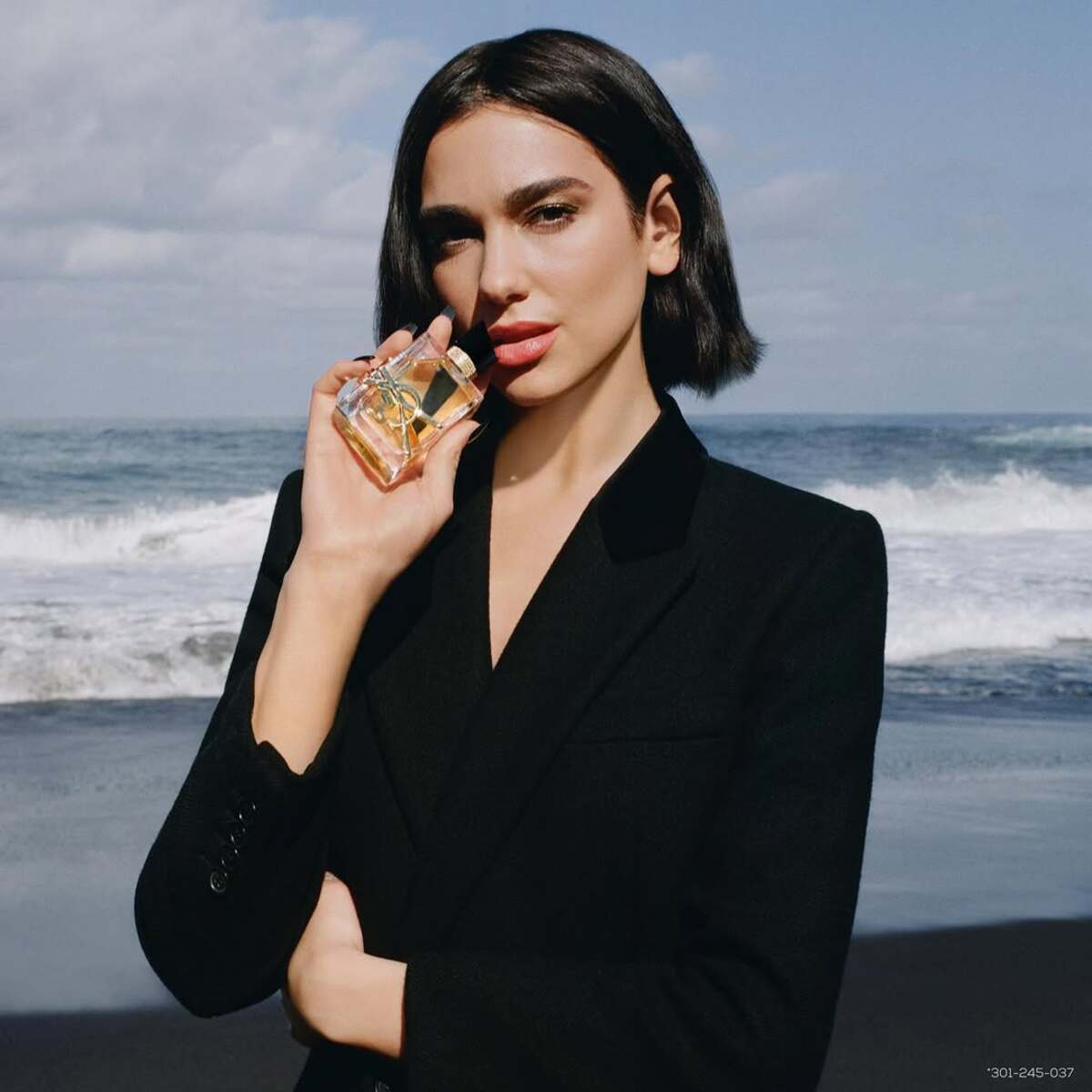 Luksusowe perfumy w Rossmannie: promocja 2025. Dua Lipa trzymająca perfumy YSL na tle morskich fal. Kobieta ma ciemne włosy do połowy szyi i nosi czarną marynarkę.