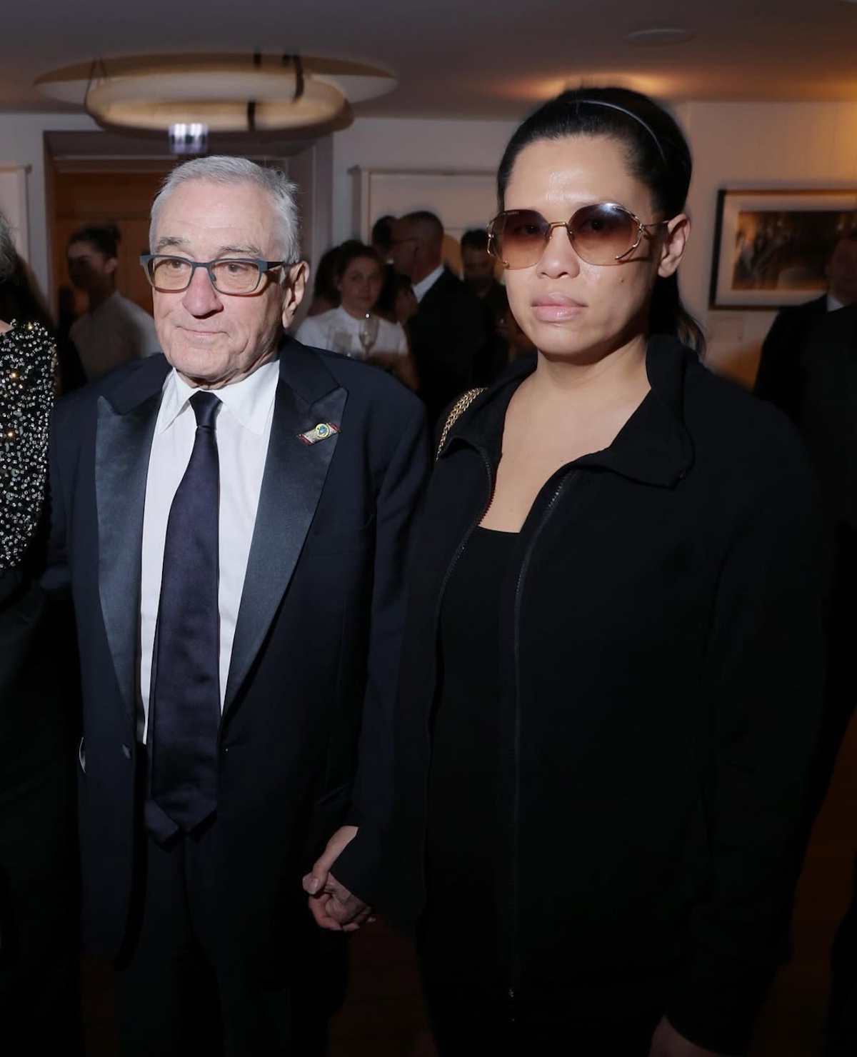 Robert De Niro ubrany w klasyczny czarny smoking z satynowymi klapami, trzymający za rękę swoją partnerkę Tiffany Chen, ubraną w czarny, sportowo-elegancki zestaw i okulary przeciwsłoneczne. Para pozuje wspólnie na ekskluzywnym evencie, otoczona przez gości w tle