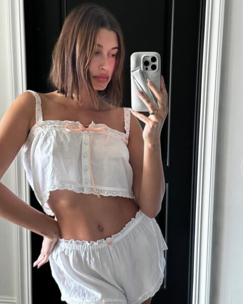 Hailey Bieber pozuje w białym komplecie piżamowym – góra jest rozpinana na guziki, ma ramiączka i różowe akcenty. Do tego nosi białe spodnie. Kobieta w krótkich włosach robi sobie selfie w lustrze.