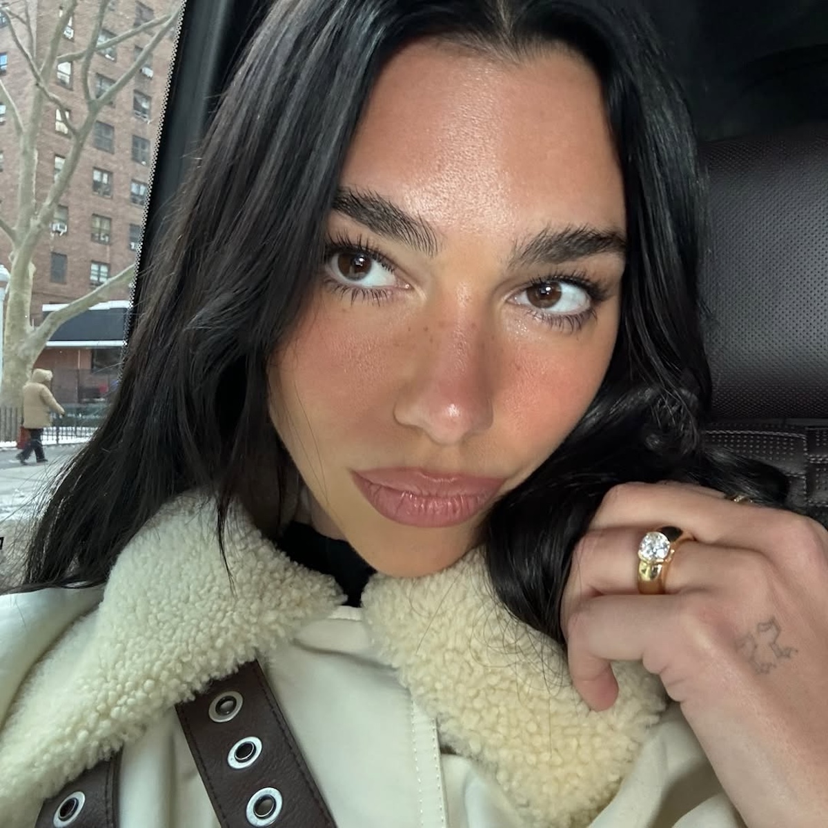 Jak dobrać kształt brwi do twarzy? Dua Lipa z gęstymi, naturalnymi brwiami pozuje w jasnej kurtce z kołnierzem z kożuchem.