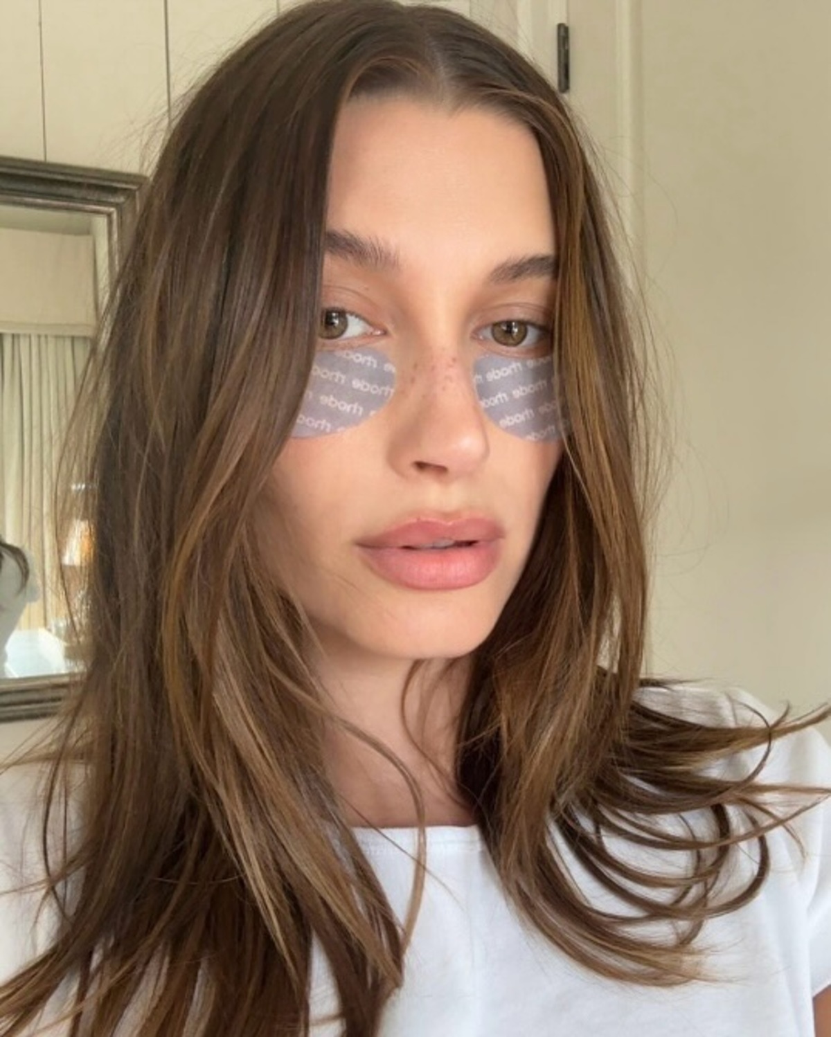 Hailey Bieber w płatkach pod oczy Rhode, które są polecane jako sposoby na cienie pod oczami. Pozuje w rozpuszczonych włosach i białej koszulce, w tle widać lustro.