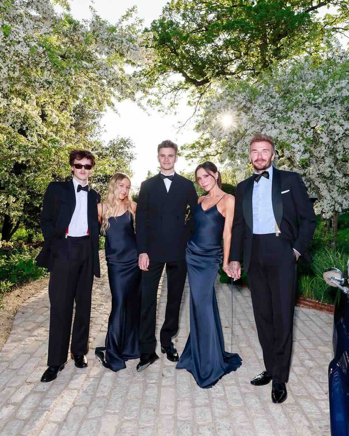 Brooklyn Beckham konflikt: Rodzina Beckhamów w wieczorowych kreacjach pozująca w ogrodzie – Grupa pięciu osób ubrana w czarno-granatowe smokingi i jedwabne suknie pozuje na tle kwitnącego ogrodu i brukowanego dziedzińca, promieniejąc elegancją i zgraniem stylizacji.