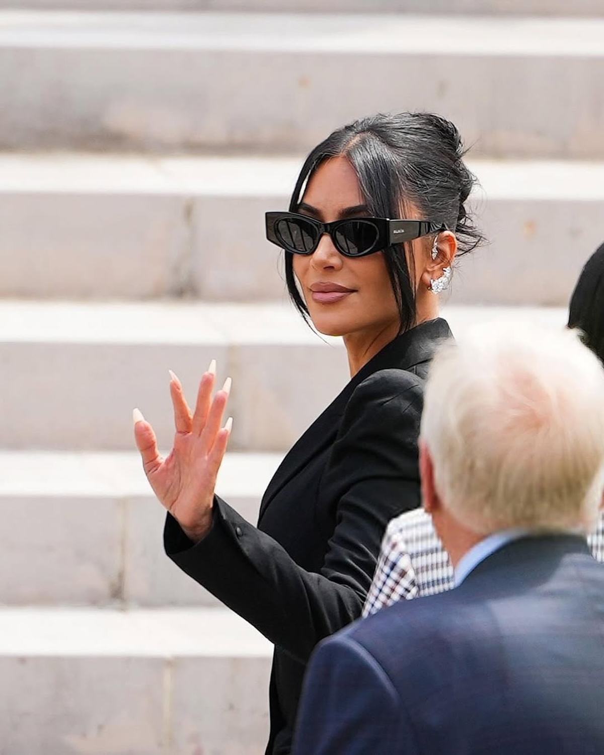 Kim Kardashian proces: Kobieta w eleganckim, czarnym garniturze i okularach przeciwsłonecznych Balenciaga macha do tłumu, eksponując długie, migdałowe paznokcie w stylu nude; ma spięte włosy i olśniewające kolczyki, stojąc na tle jasnych schodów.