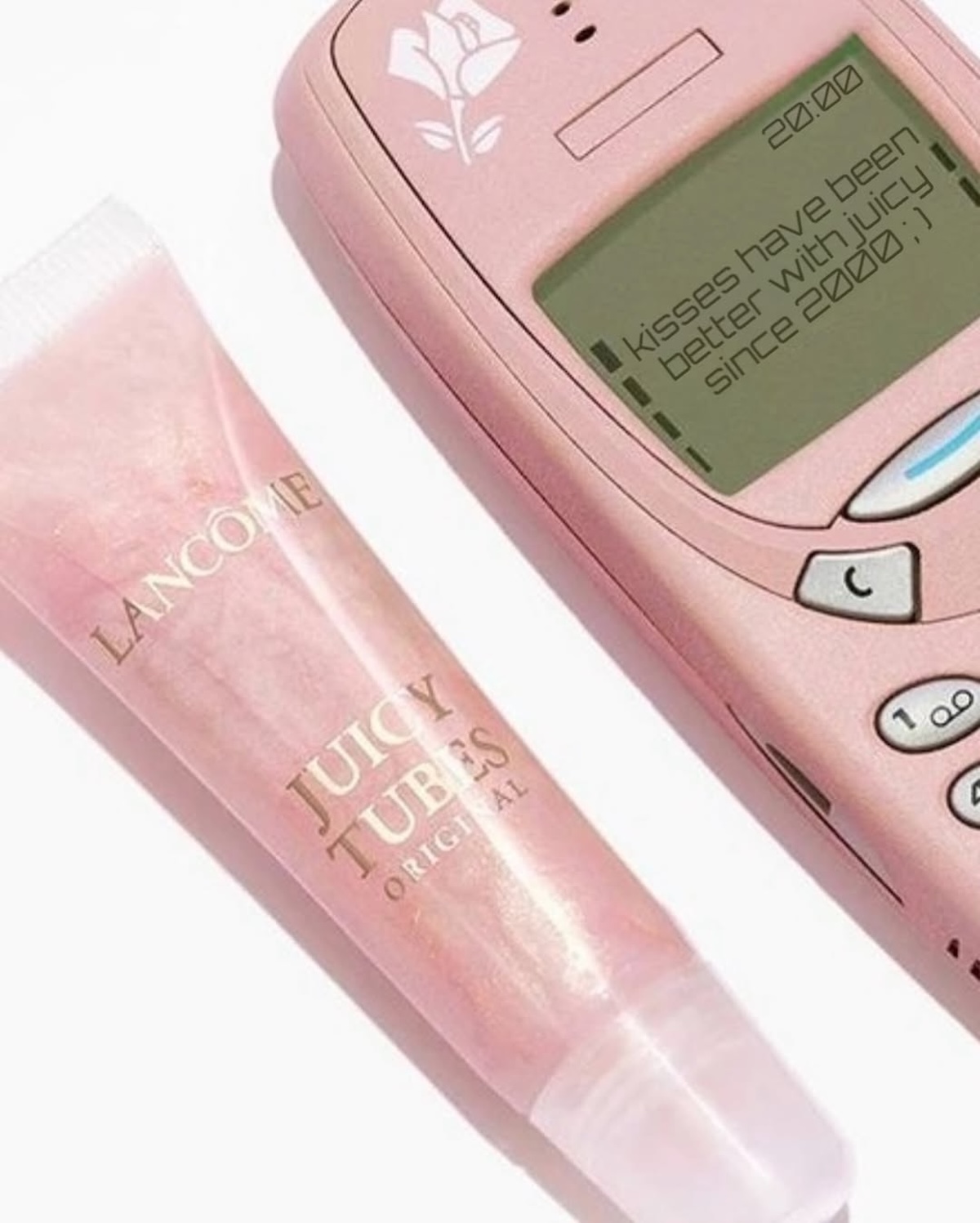 Lancôme Juicy Tubes: Lancôme Juicy Tubes zestawiony z różowym telefonem komórkowym typu retro Nokia z logo róży i napisem na ekranie – „Kisses have been better with Juicy since 2000 ;)”, obrazek inspirowany stylistyką lat 2000., podkreślający kultowy status produktu.