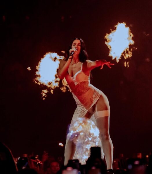 Dua Lipa Ewa Farna: Dua Lipa w koronkowej, przezroczystej sukni podczas spektakularnego występu z płomieniami na scenie. Artystka śpiewa i gestykuluje, stojąc na tle buchającego ognia i tłumu z telefonami.