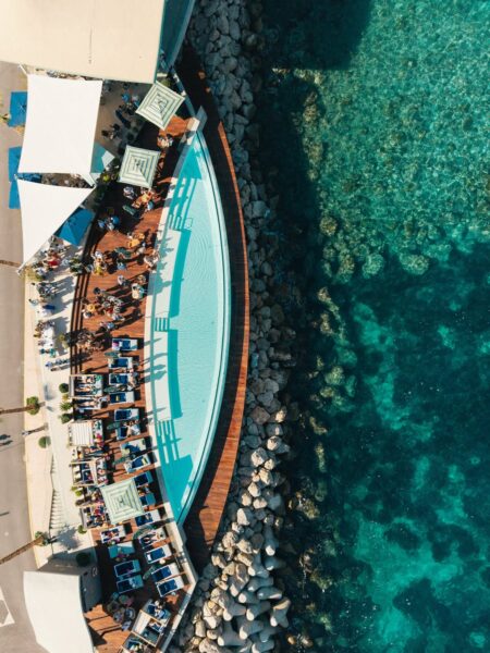 Zdjęcie z lotu ptaka przedstawiające elegancki klub plażowy z tarasem wzdłuż brzegu Morza Śródziemnego. Na drewnianym pokładzie wokół półkolistego basenu rozmieszczone są leżaki, stoliki i parasole, przy których wypoczywają goście. Po prawej stronie widać przejrzystą, turkusową wodę i kamieniste dno morza. Całość utrzymana w słonecznej, wakacyjnej atmosferze z widokiem na błękitne wybrzeże.