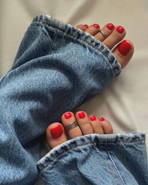 Zdjęcie stóp z pedicure w kolorze czerwonym. Na palcach znajdują się pierścionki. Widać jeansowe nogawki. Nogi pozują na tle beżowego materiału.