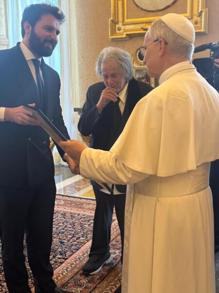 W eleganckim, bogato zdobionym wnętrzu z pozłacanymi ramami obrazów i wzorzystym dywanem papież ubrany w białą sutannę rozmawia z dwoma mężczyznami. Jeden z nich, młodszy, trzyma w rękach dokument i uśmiecha się, drugi, starszy z długimi siwymi włosami, sprawia wrażenie zamyślonego. Scena ukazuje formalne, ale serdeczne spotkanie w Watykanie.