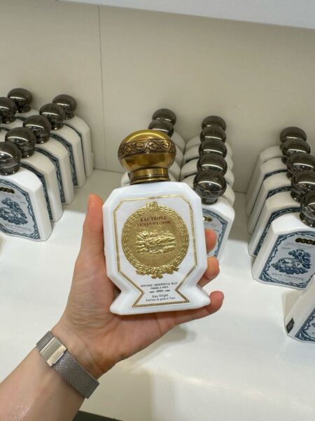 Elegancki biały flakon perfum Eau Triple Lichen d’Écosse od Officine Universelle Buly, inspirowany XVIII-wieczną estetyką apteczną. Złota plakietka i zdobna zakrętka tworzą wyjątkowy design, który łączy luksus z historią francuskiej perfumerii.