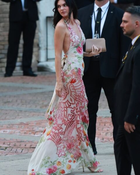 Zdjęcie celebrytki Kendall Jenner na ślubie Jeffa Bezosa i Lauren Sanchez. Ma na sobie długą, biało-czerwoną sukienkę w kwiaty. Ma zielone sandałki i nude pedicure, a w ręce beżową torebkę. Przechodzi przez plac i ogląda się za siebie. Otaczają ją pracownicy ochrony ubrani w garnitury.