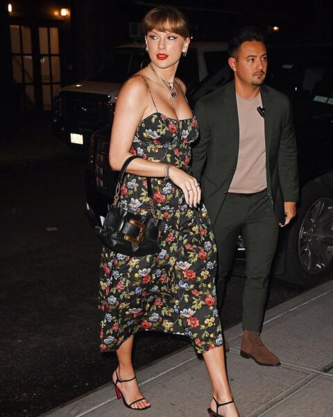 Zdjęcie piosenkarki Taylor Swift. Idzie przez chodnik, ulicą, wieczorem. Ubrana jest w czarną sukienkę rozkloszowaną w kwiaty, na ramiączkach, midi. Na nogach ma czarne sandałki i czerwony pedicure. Na ramieniu czarna torebka. Włosy spięte, ma tylko blond grzywkę na czole.