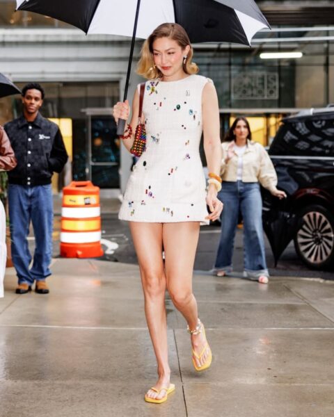Zdjęcie modelki Gigi Hadid w krótkiej, białej sukience bez rękawów, z drobnymi, kolorowymi zdobieniami. Ma pomarańczowe klapki na nogach i zielone paznokcie u stóp. Na ramieniu ma kolorową, plecioną torebkę. Włosy blond, ścięte na boba. Ma na sobie dużo biżuterii, w tym bransoletki i kolczyki. Niesie parasol i idzie ulicą.