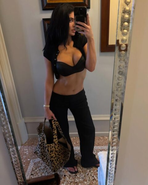 Zdjęcie celebrytki Kylie Jenner ubranej w czarne spodnie i czarny stanik. Ma w ręce dużą torebkę ze wzorem panterki. Ma czarne, rozpuszczone włosy. Robi sobie selfie telefonem w lustrze na korytarzu eleganckiego hotelu.