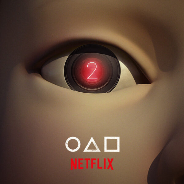 Zbliżenie na mechaniczne oko kultowej lalki z „Squid Game”, z cyfrową cyfrą 2 w centrum — zapowiedź 2. sezonu serialu Netflix z charakterystycznymi symbolami koła, trójkąta i kwadratu pod logo.