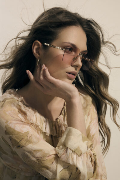 Kobieta w delikatnych różowych okularach przeciwsłonecznych Gucci z perłowymi zausznikami, w romantycznej bluzce w kwiaty – elegancka i eteryczna propozycja w stylu soft glam.