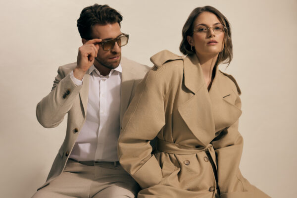 Para – mężczyzna i kobieta – w beżowych total lookach z okularami korekcyjnymi i przeciwsłonecznymi, wpisującymi się w minimalistyczną elegancję. Mężczyzna w geometrycznych oprawkach, kobieta w subtelnych okularach vintage.