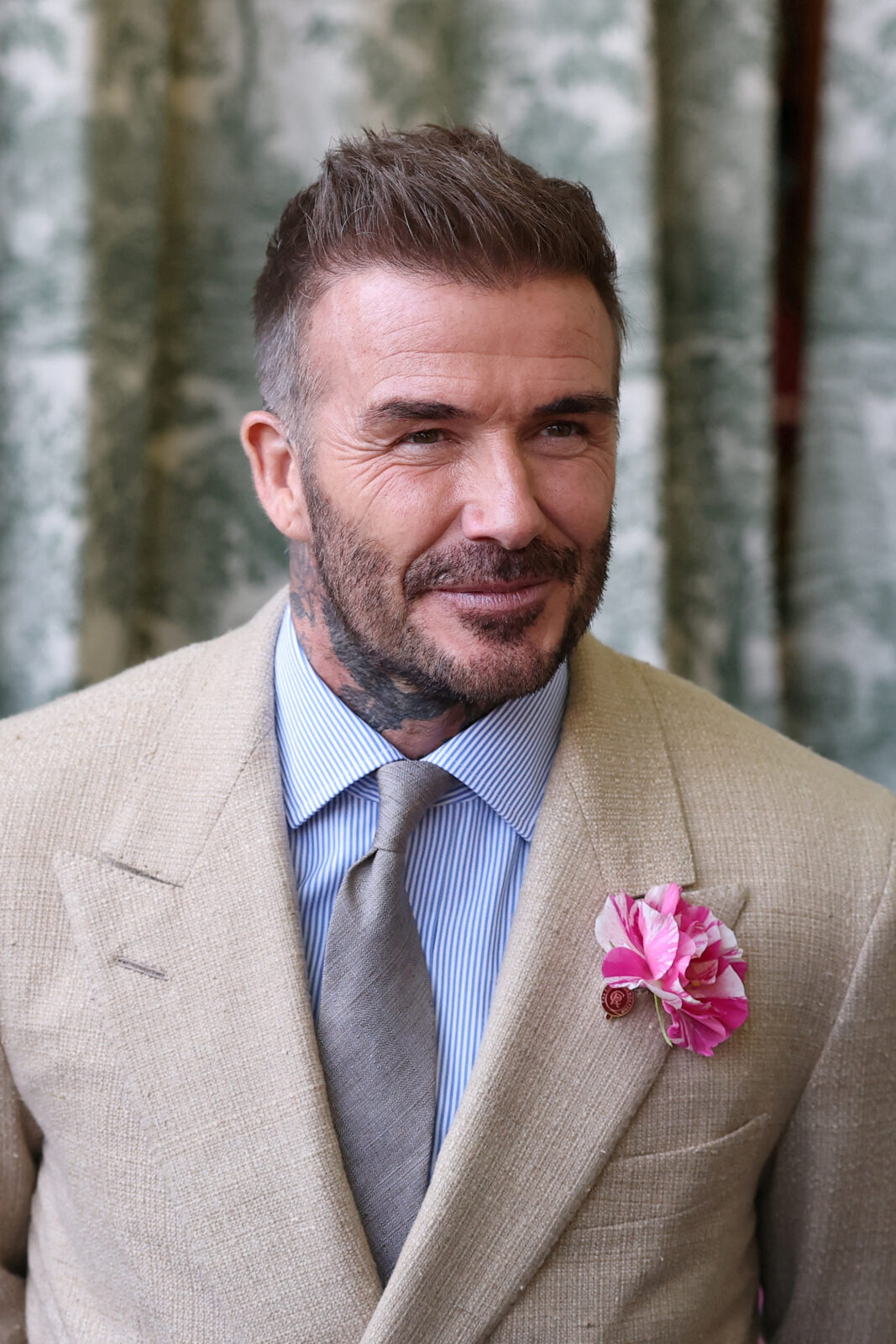 David Beckham tytuł szlachecki: ubrany w beżową, teksturowaną marynarkę dwurzędową i szarą krawatkę, z niebieską koszulą w paski. Do klapy marynarki przypięty ma różowo-biały kwiat ozdobny. Beckham patrzy lekko w bok i uśmiecha się, stojąc na tle zielonej tapety. Jego stylizacja emanuje elegancją w stylu angielskiego dżentelmena.