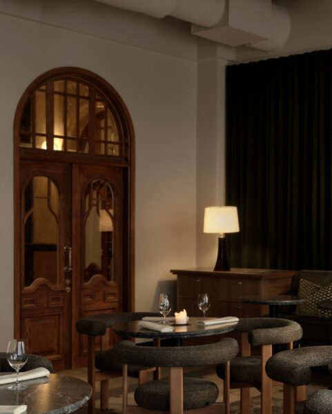 Eleganckie, przytulne wnętrze restauracji z przyciemnionym oświetleniem. Na pierwszym planie okrągłe stoliki z ciemnego kamienia, nakryte białymi serwetkami, kieliszkami i świecami. W tle drewniane drzwi z łukowym zwieńczeniem i przeszkleniami oraz komoda z lampą stołową. Klimat wnętrza podkreślają ciemnozielone zasłony i miękkie, tapicerowane krzesła w odcieniach szarości i drewna. Całość emanuje ciepłem i elegancją w stylu klasycznej nowoczesności.
