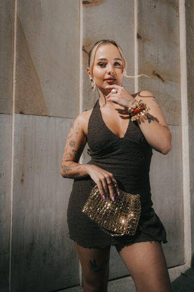 Martyna Pysia w eleganckiej, brązowej mini sukience bez ramiączek, trzymająca złotą błyszczącą kopertówkę. Ma na sobie duże kolczyki, kilka pierścionków i bransoletki. Ujęcie wykonane w mocnym świetle dziennym, z betonową ścianą w tle, podkreśla nowoczesny glamour i odważny styl.