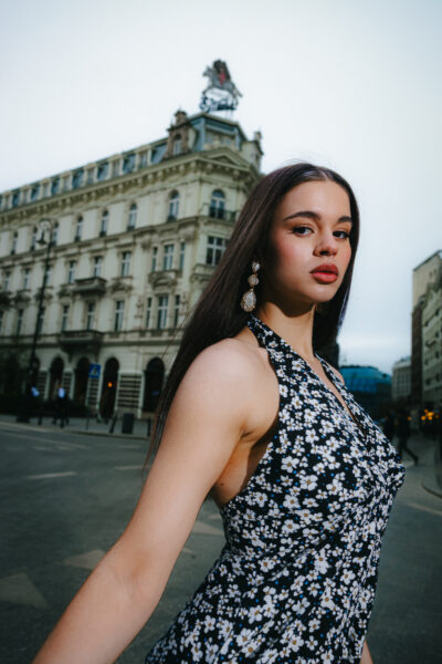 Zbliżenie na Anastasiię Zakhandrevych w letniej sukience w kwiaty i dużych złotych kolczykach, sfotografowaną na tle architektury warszawskiego Śródmieścia. Stylizacja inspirowana miejskim romantyzmem z nutą retro.