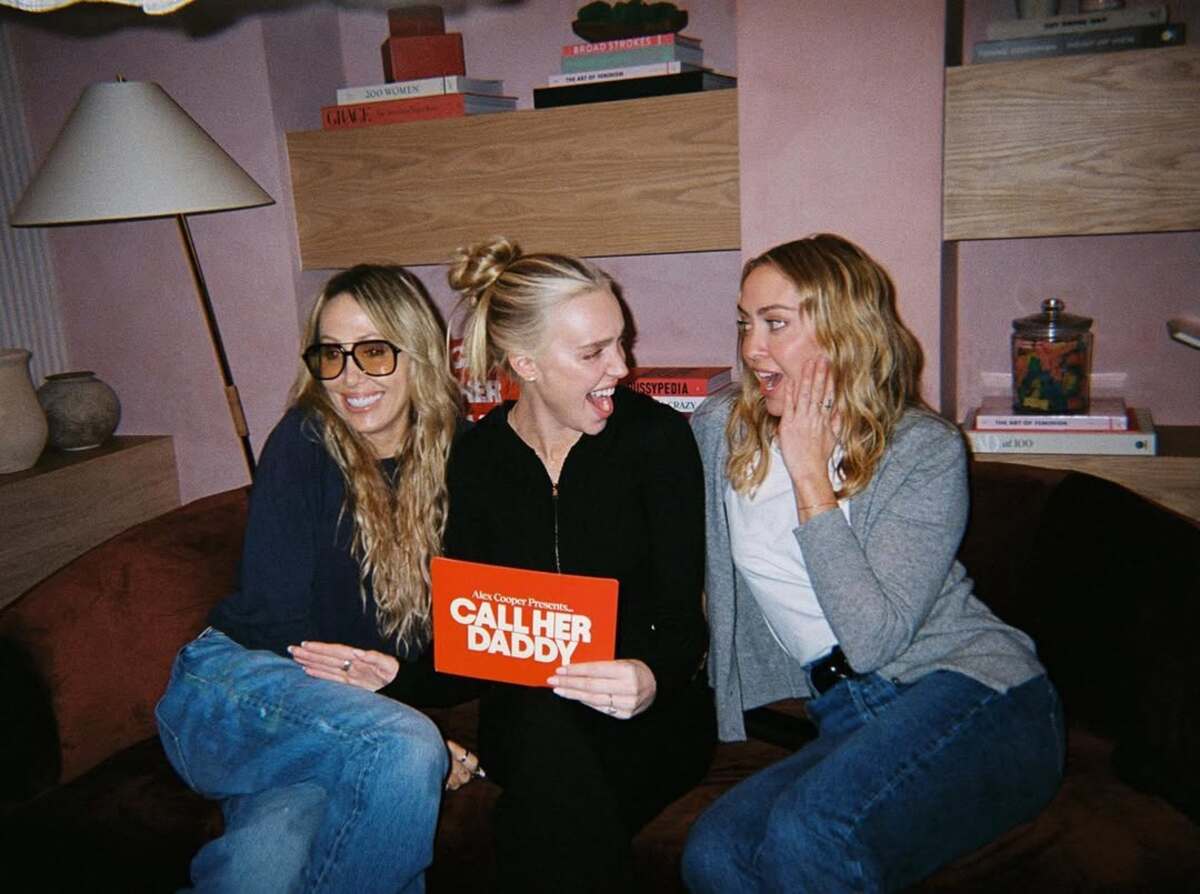 Najlepsze podcasty gwiazd. Zdjęcie prowadzącej podcast Call Her Daddy z dwiema kobietami – po lewej nosi okulary przeciwsłoneczne, ciemną bluzkę i jeansy, a po prawej kobieta ma na sobie szarą marynarkę, białą bluzkę i jeansy. Autorka podcastu jest w czarnym total looku.