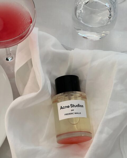Flakon perfum Acne Studios par Frédéric Malle ustawiony na białej, lekko pogniecionej tkaninie, obok kieliszka z różowym napojem oraz szklanki z wodą. Perfumy mają prostą, przezroczystą butelkę z czarnym korkiem i minimalistyczną etykietą. Kompozycja nawiązuje do estetyki francuskiego stołu, łącząc nowoczesny design z subtelną elegancją.
