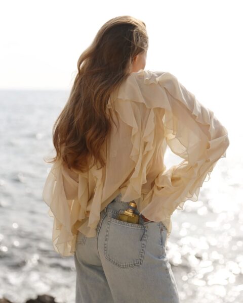 Zdjęcie kobiety stojącej tyłem w długich rozpuszczonych włosach na tle morza. Kobieta nosi jasną bluzkę z falbankami ze zwiewnego materiały i jeansy. Z kieszeni spodni wystaje flakon perfum ze złotą nakrętką.